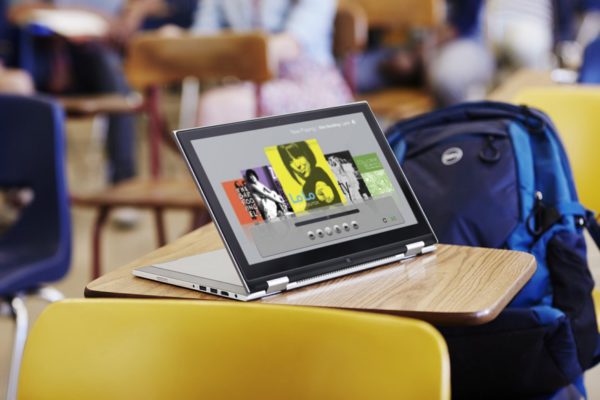 Dell Inspiron 11 3000 Series 2-in-1 Touch ( Model 3147 ) notebook computer in tablet position ( with keyboard serving as stand ), on a student desk in a classroom, with Dell Energy backpack in seat and blurry students in background.